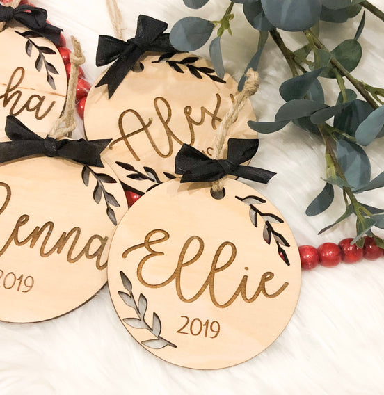 Personalized round wooden Christmas ornaments with engraved names, leafy borders and black bows.