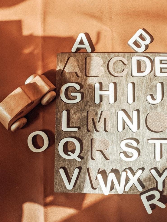 Close-up of the alphabet puzzle's letter cutouts and pale wooden pieces beside a toy car.