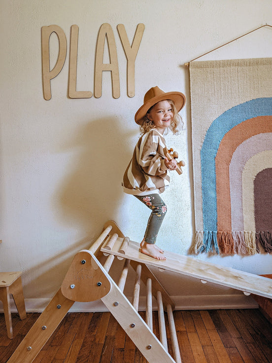 Wooden PLAY letters above a child on a climbing triangle beside a rainbow wall hanging.