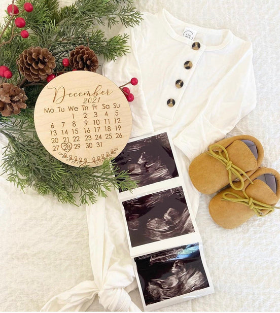 December wooden calendar pregnancy-announcement disc with ultrasound photos, baby clothing and shoes among evergreen branches.