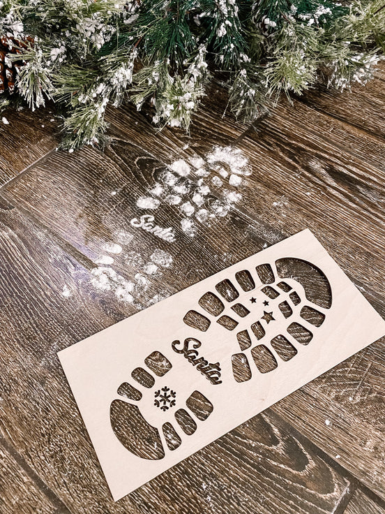 Close-up of a wooden boot-print stencil and white powder footprint beside a frosted Christmas tree.