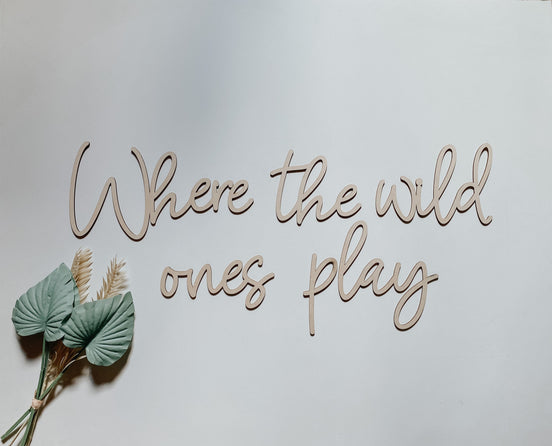 Natural wooden Where the Wild Ones Play script lettering on a wall beside green decorative leaves.