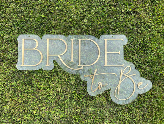 Clear acrylic Bride to Be sign with gold lettering lying on grass.