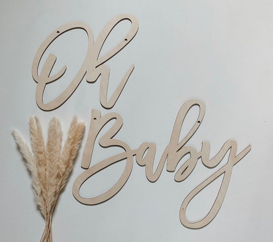 Natural wooden Oh Baby script lettering against a pale wall beside dried grass.