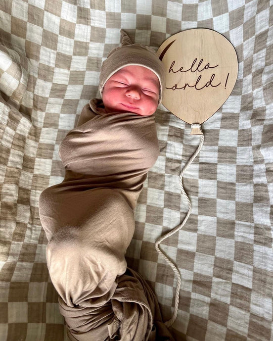 Swaddled newborn beside an engraved Hello World balloon-shaped wooden plaque.