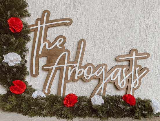 The Arbogasts family sign with raised white script on a woodgrain backing, beside holiday greenery.