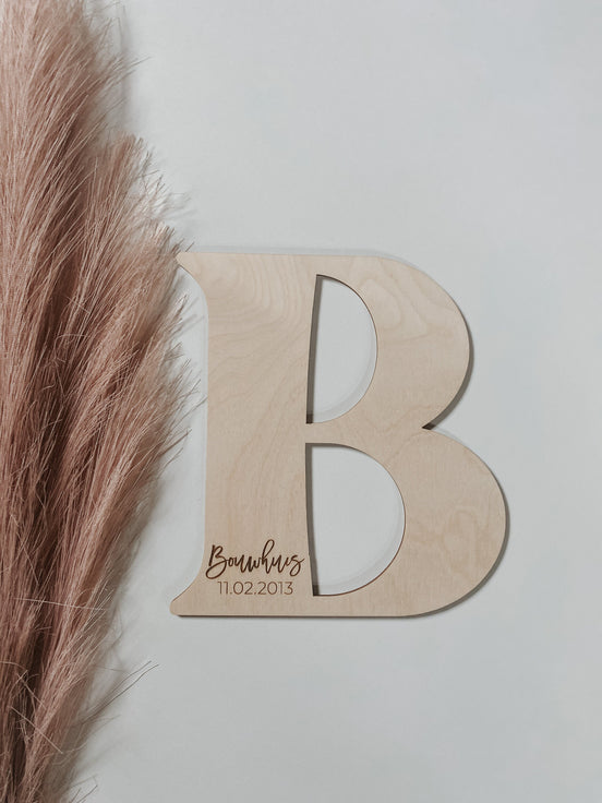 Natural wooden B wedding guest-book letter with an engraved surname and date on a pale wall.