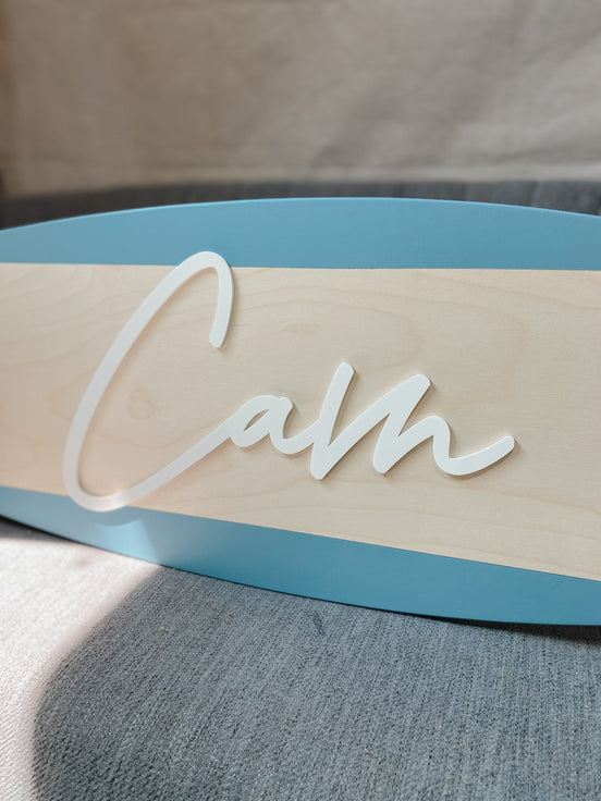 Close-up of raised Cam script and natural wood grain on a blue-bordered surfboard sign.