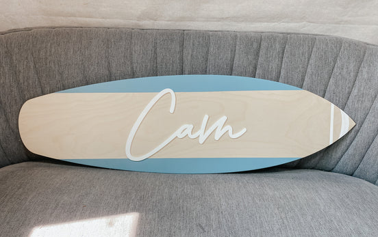 Blue-edged wooden surfboard name sign with raised white Cam lettering on a gray chair.