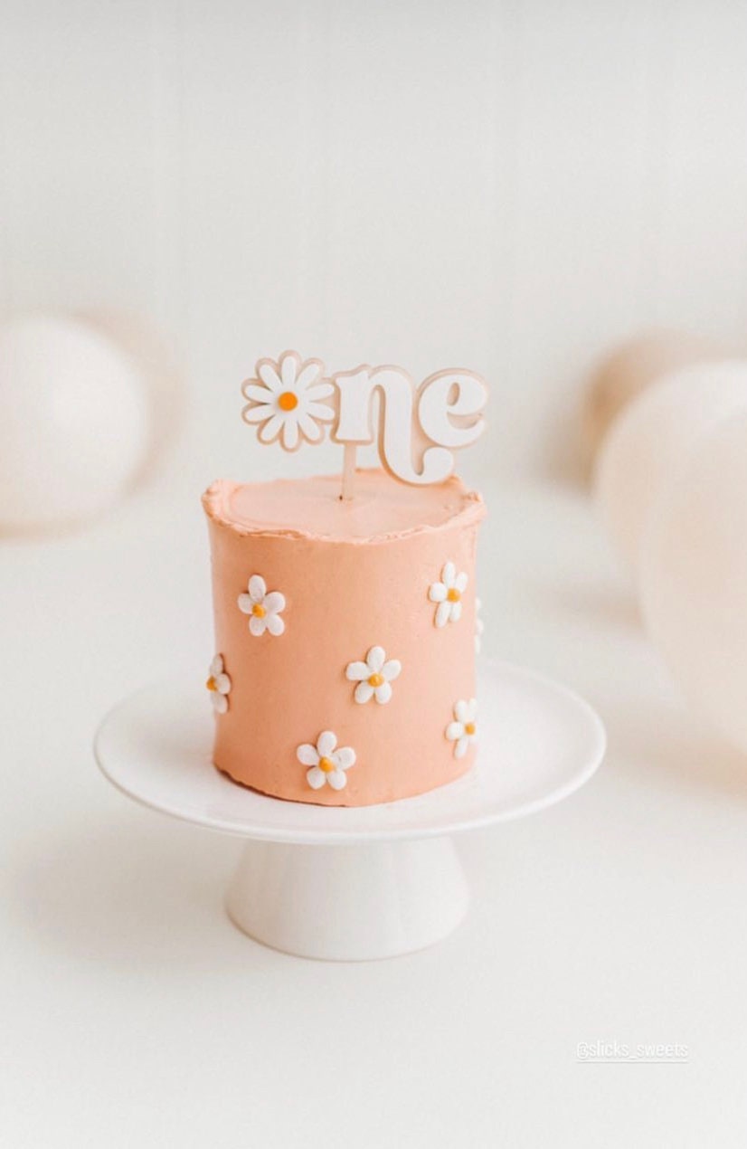 White One cake topper with a daisy-shaped O on a peach cake decorated with daisies.