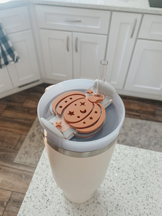 Pumpkin topper with celestial cutouts fitted around a clear straw on a cream tumbler.