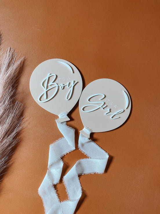 Boy and Girl balloon announcement plaques with raised white script and white ribbons on a rust background.
