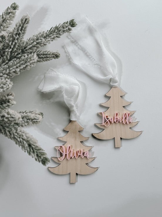 Two natural-colored Christmas tree name tags with pink script names and white ribbon loops.