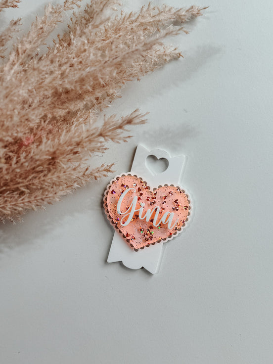 Pink Olivia heart topper with raised white script and a white arrow-shaped base beside dried grasses.