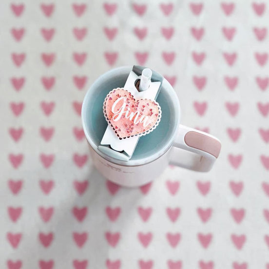 Olivia heart-shaped personalized tumbler topper fitted on a cup lid against a pink heart background.