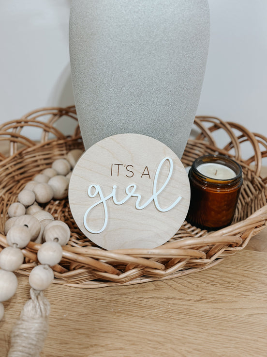 Round wooden It's a Girl announcement sign with raised white script beside beads and an amber jar.