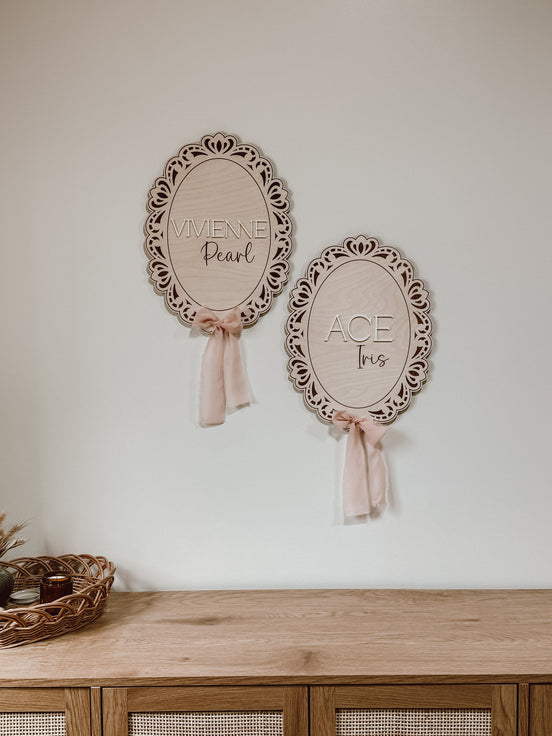 Vivienne Pearl and Ace Ives ornate oval wooden name signs with pale ribbon bows above a dresser.