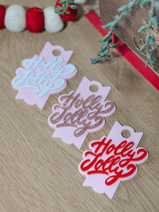 Three pink Holly Jolly toppers with pale, glitter and red lettering lying on wood.