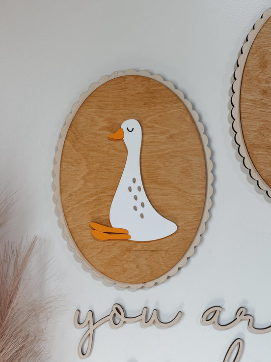 Close-up of a white goose with orange feet on a scalloped oval wooden wall plaque.