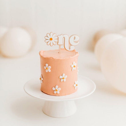 Close view of a daisy One topper on a small peach first-birthday cake.