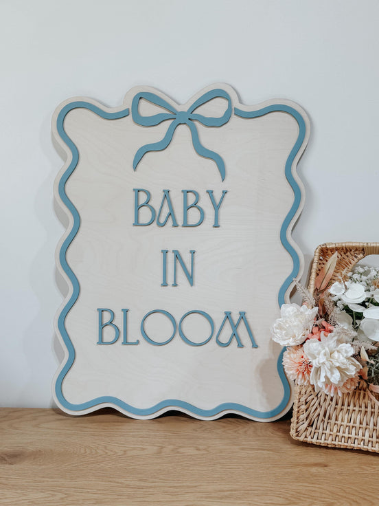 Baby in Bloom wooden sign with a blue wavy border and bow, standing beside a flower basket.