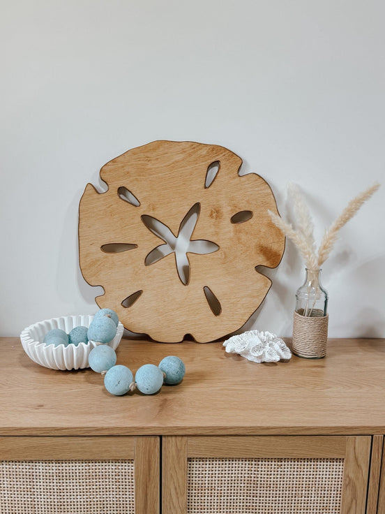 Large wooden sand-dollar decoration on a dresser with blue felt beads, white coral and dried grasses.