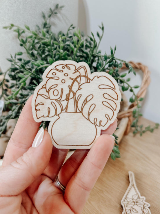 Hand holding a wooden monstera plant magnet with engraved leaves and a white pot outline.