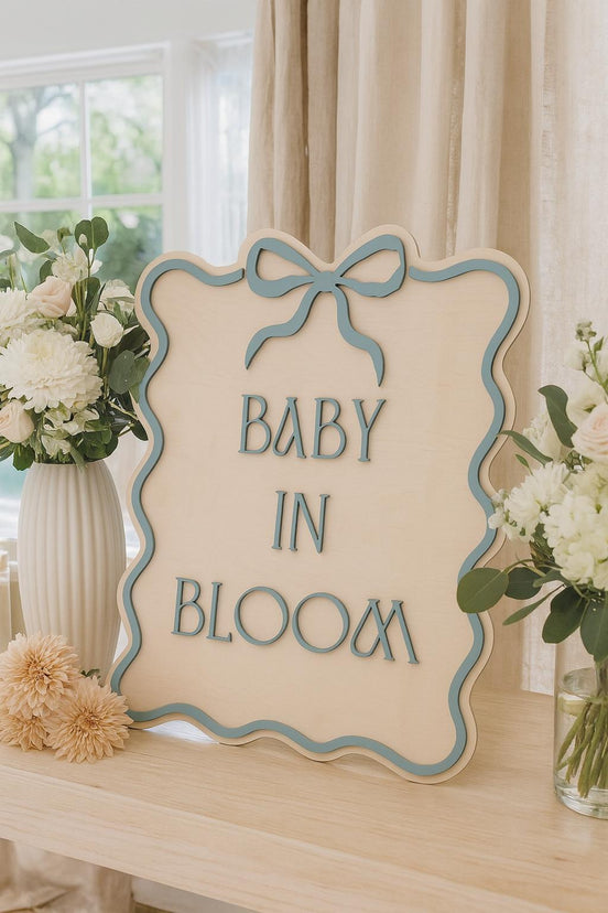 Blue-bordered Baby in Bloom sign with a bow, displayed among white flowers.