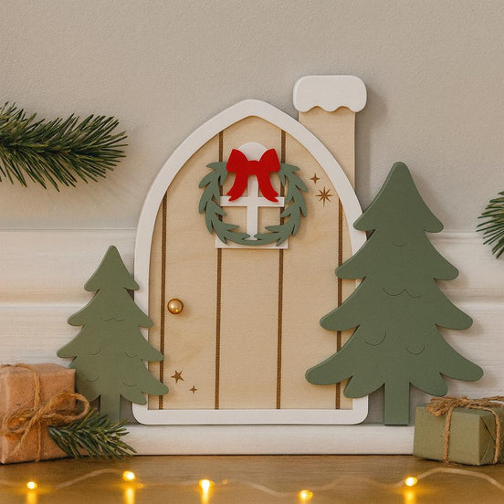 Miniature arched elf door with a wreath, red bow, snowy chimney and green trees.