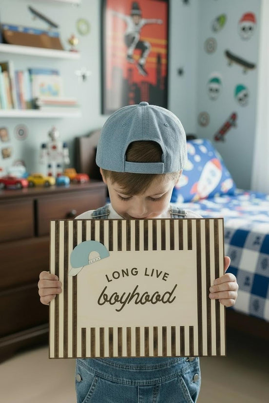 Child holding a framed Long Live Boyhood sign with vertical wood slats and a small blue cap accent.