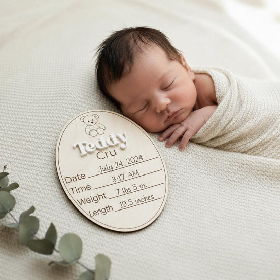 Sleeping newborn beside a personalized Teddy Cru wooden birth-announcement plaque.