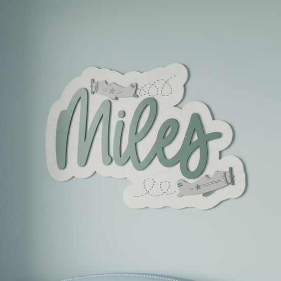 Miles airplane-themed name sign with raised sage script, engraved flight paths and cloud shapes.