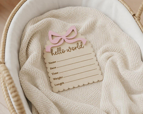 Scalloped wooden Hello World birth announcement plaque with a pink bow on a knitted blanket.